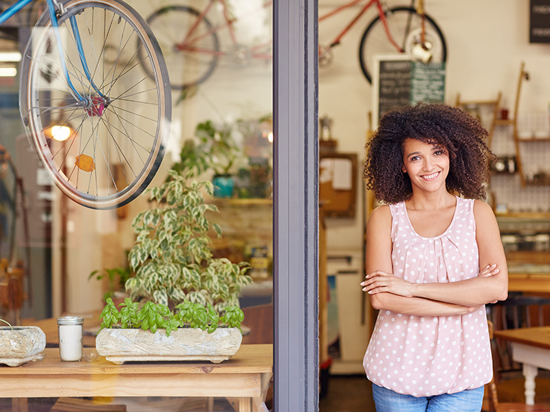 Young mixed race woman smiling, while standing in the door of her cafe with her arms folded proud to be the owner of a small business