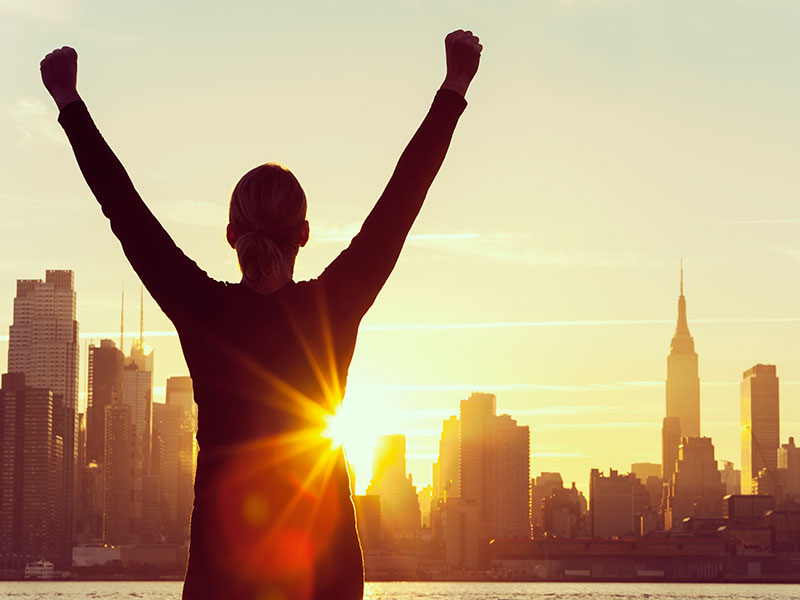 silhouette of a successful woman or girl arms raised celebrating at sunrise or sunset in front of the New York City Skyline