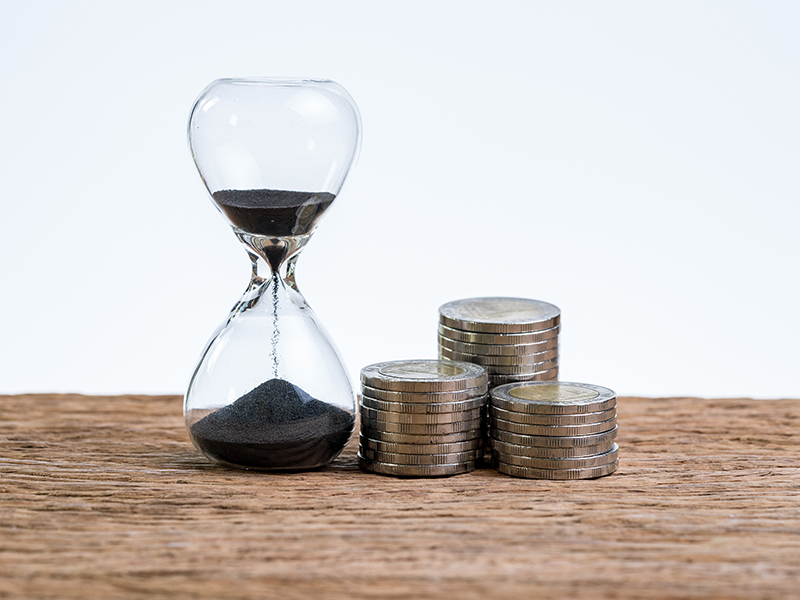 Financial or investment time counting with hourglass or sandglass and stack of coins on wooden table with white background.