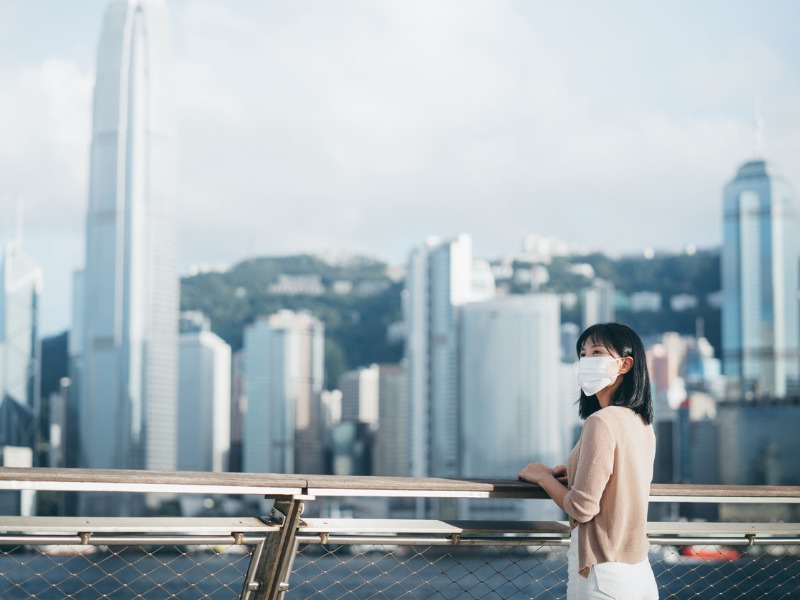 Woman wearing a protective face mask to protect and prevent from the spread of viruses in the city, looking away while standing by the promenade of Victoria harbour against Hong Kong city skyline