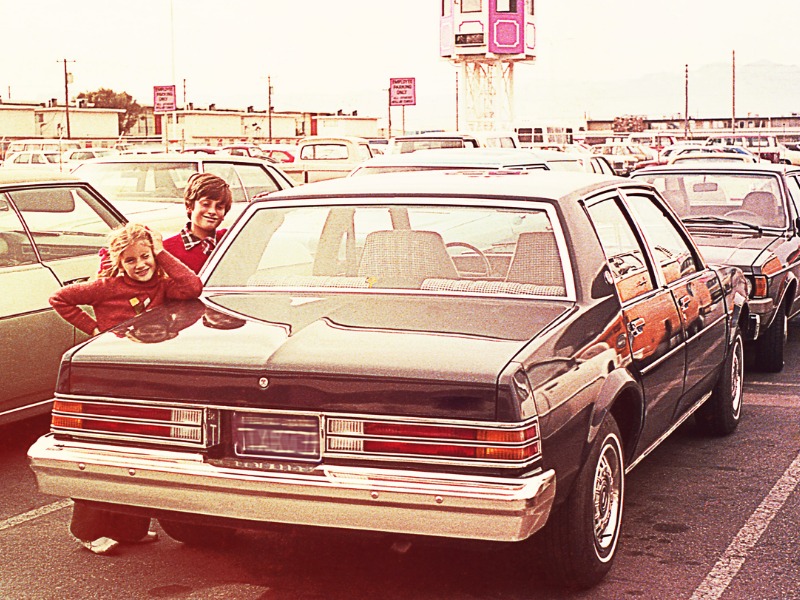 Vintage image of a children standing beside a car in a parking lot.