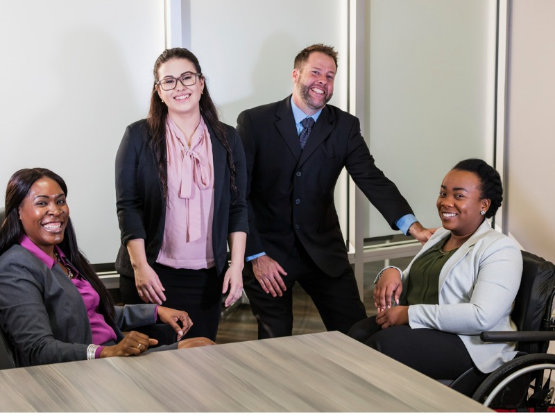 A group of four multi-ethnic business people meeting in an office boardroom. The man, wearing a suit, and three women are smiling at the camera. The young woman standing is Hispanic. One of the African-American women is in a wheelchair. Her physical disability is due to spina bifida. They are looking at the camera.