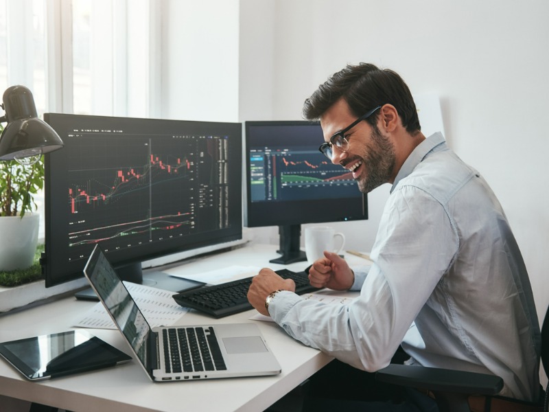 Happy young businessman or trader in formalwear and eyeglasses using laptop and smiling while sitting in his modern office. Stock exchange. Financial trading concept. Investment concept