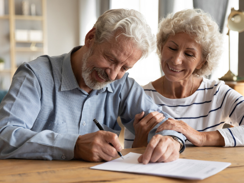 Happy mature retired couple signing a document around the kitchen table|Wealth by HomeEquity Bank logo